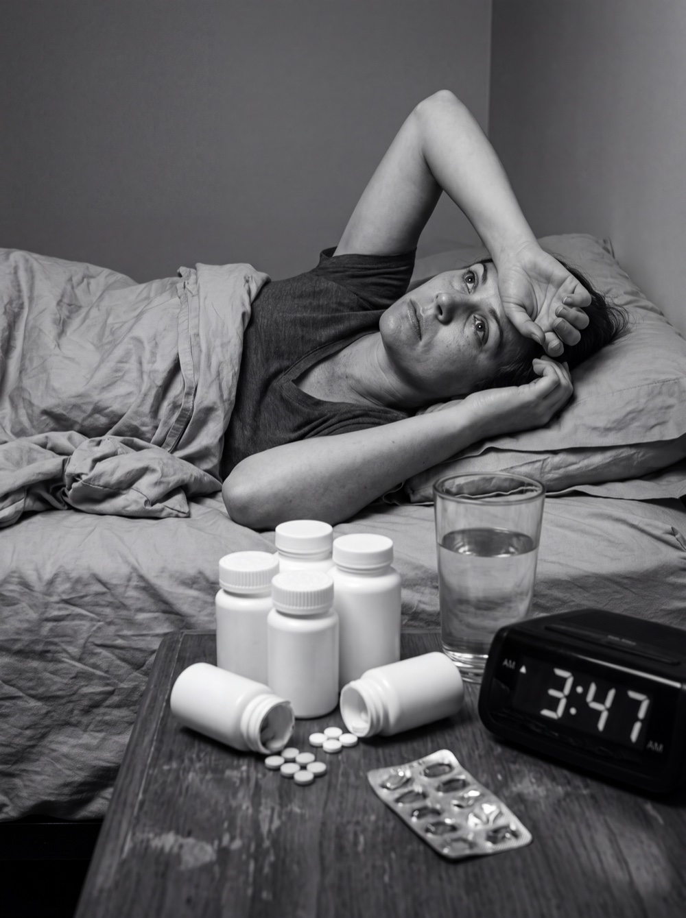 A woman lying awake at 3:47am beside a nightstand of failed over-the-counter sleep aids